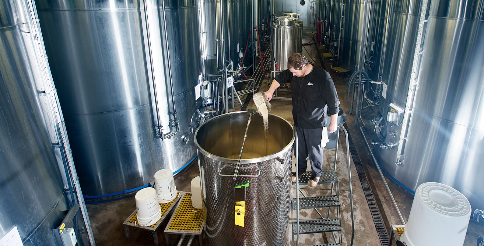 A picture showing the cider making process and large brewing tanks. A man is adding yeast to water in a smaller tank